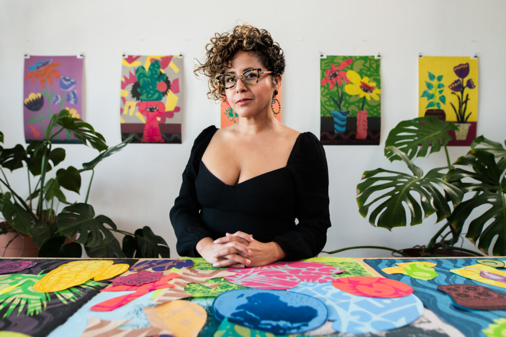 Favianna stands at a work desk with posters behind her and colorful cutout shapes in front